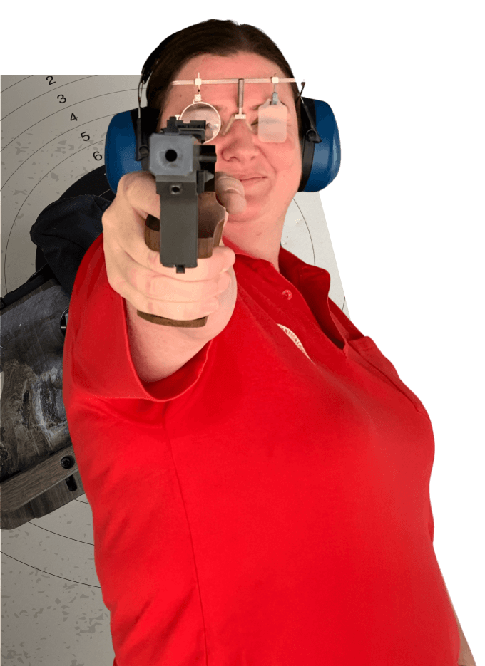 Person in red shirt aiming a handgun with protective glasses and ear protection, target in background.